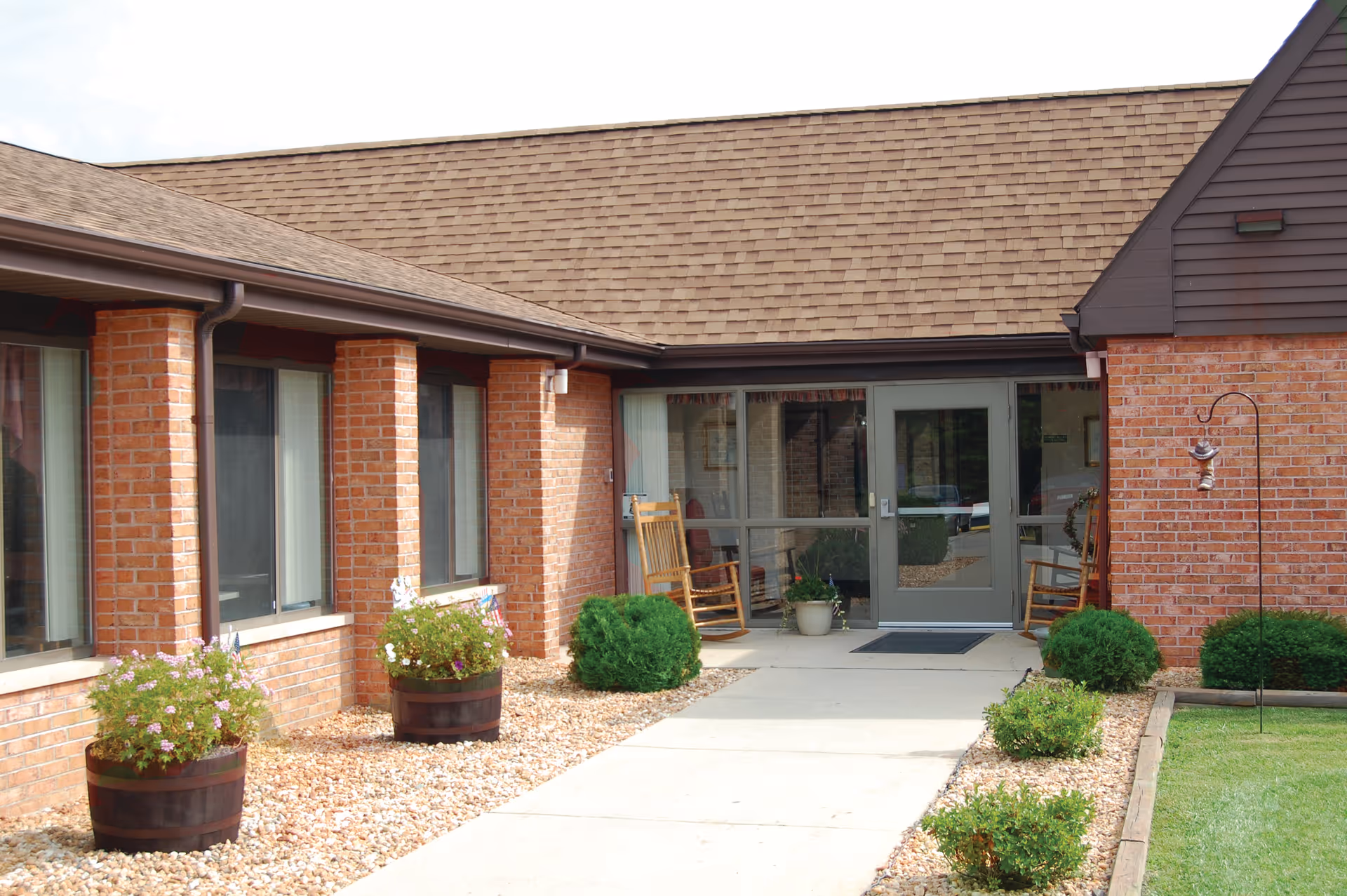 Exterior view of a brick building with a walkway leading to a glass door entrance. There are two wooden rocking chairs on the porch, potted plants, and small bushes along the walkway.