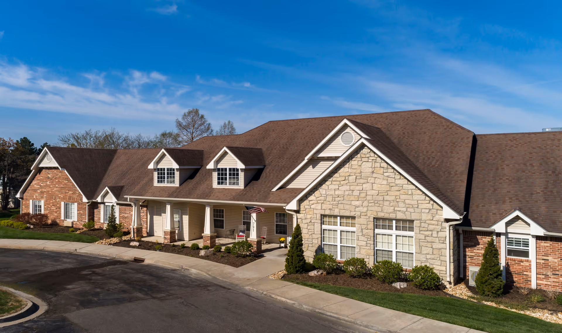 Exterior view of a single-story senior living facility building with a combination of brick and stone facade, multiple windows, a brown shingled roof, and a small covered porch with an American flag. The building is surrounded by landscaped bushes and a paved driveway under a clear blue sky.