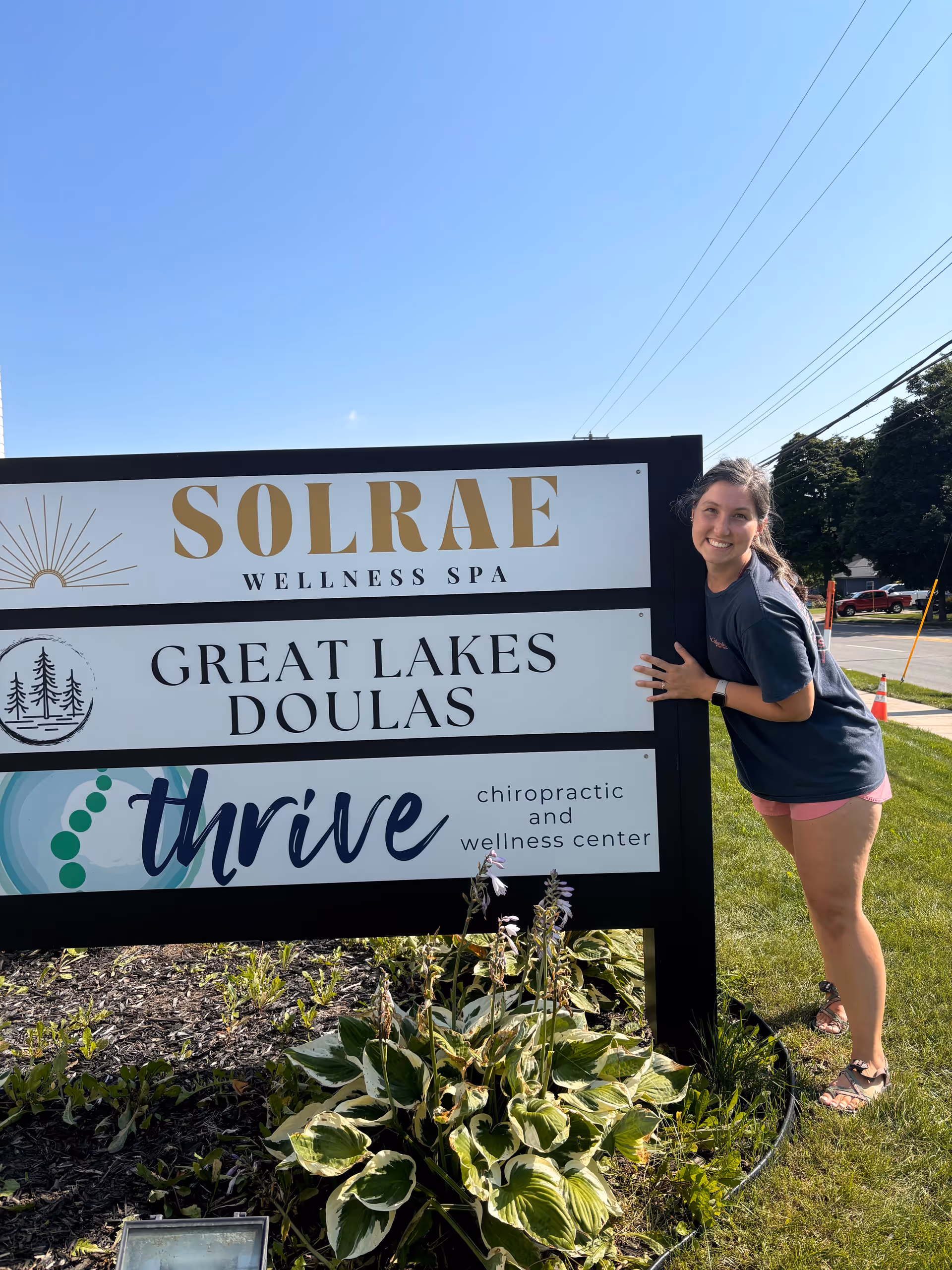 Smiling woman stands beside a large exterior sign listing wellness businesses.