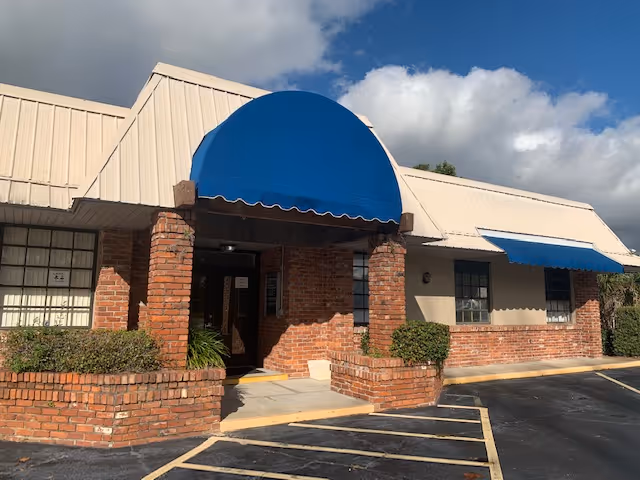 Senior living facility entrance with blue awning, brick facade, and a paved parking lot.