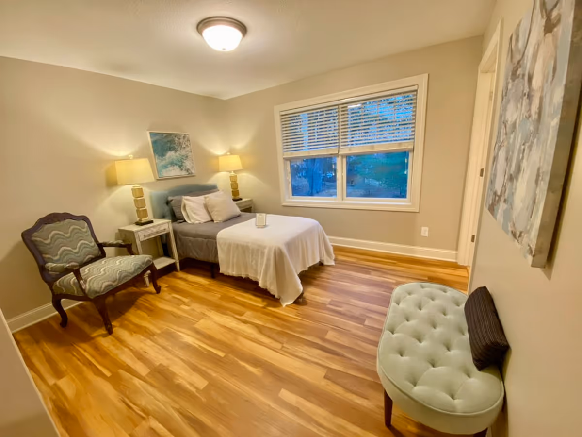 Cozy senior living bedroom featuring a bed, chairs, lamps, window, and ottoman.