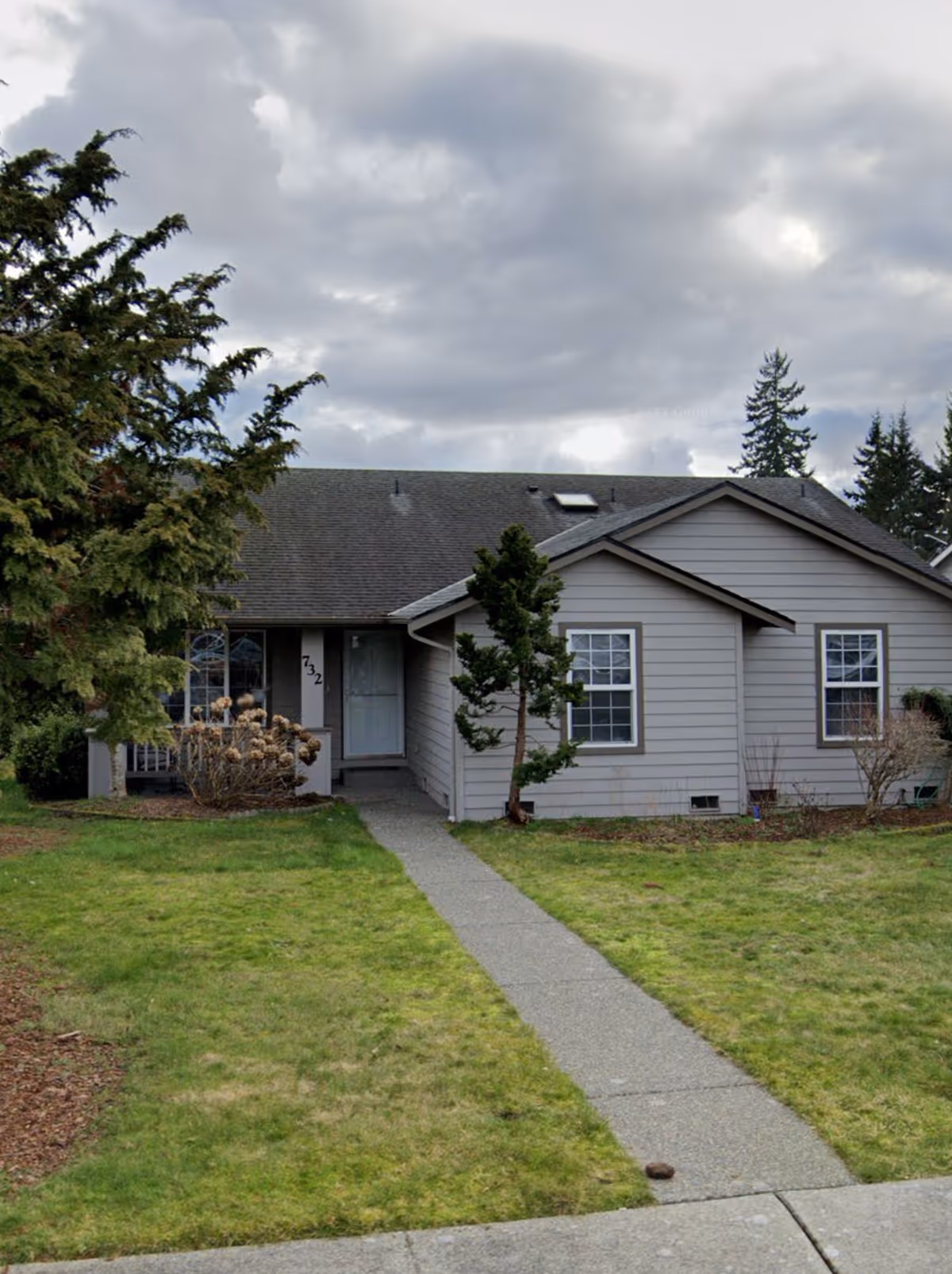 A single-story gray home with a concrete front path, lawn, and trees under cloudy sky.