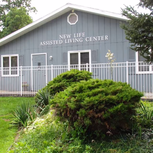 Exterior view of a gray building with white trim labeled 'New Life Assisted Living Center' with a white fence in front and green bushes and grass in the foreground.