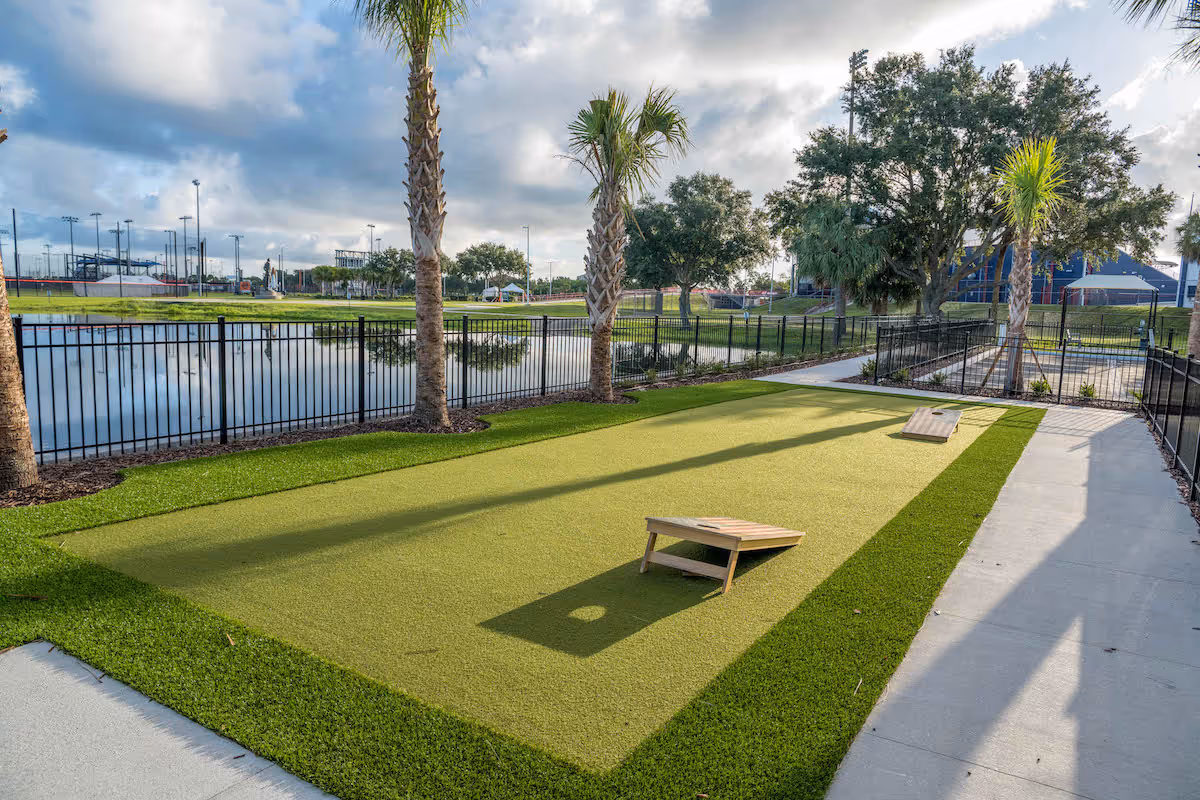 Outdoor bocce ball court with two wooden bocce ball ramps on artificial grass, surrounded by palm trees and a black metal fence. In the background, there is a pond, trees, and a sports field under a partly cloudy sky.