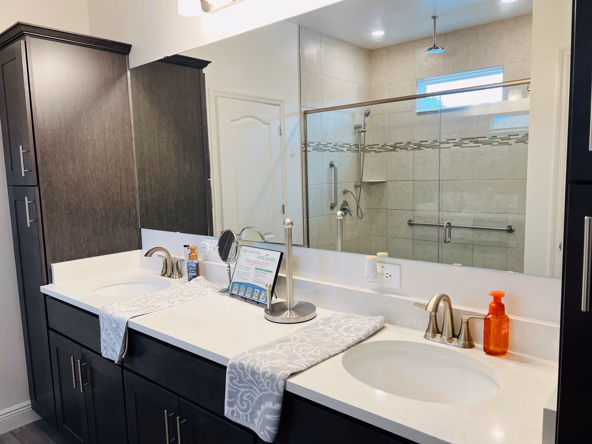 A modern bathroom with a double sink vanity featuring dark wood cabinets and white countertops. Each sink has a silver faucet and a patterned hand towel draped over the edge. On the countertop, there is a small mirror, a soap dispenser, and a framed informational sign. A large mirror spans the length of the vanity, reflecting a tiled walk-in shower with grab bars and a rainfall showerhead. The bathroom has a clean and accessible design.