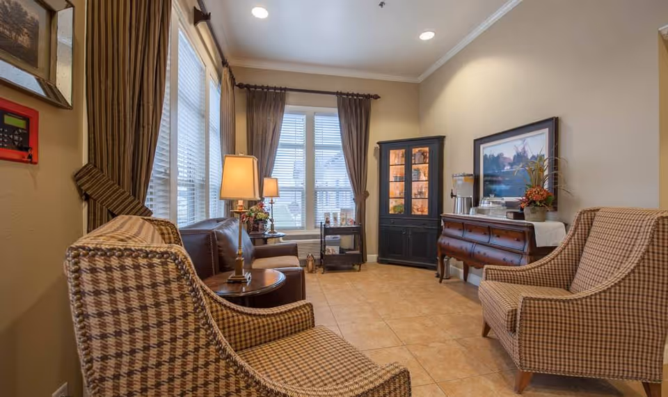A cozy senior living common area with two patterned armchairs, a brown leather sofa, two table lamps, a black display cabinet, a wooden sideboard with a beverage dispenser, and a framed landscape painting on the wall. Large windows with brown curtains allow natural light into the room.