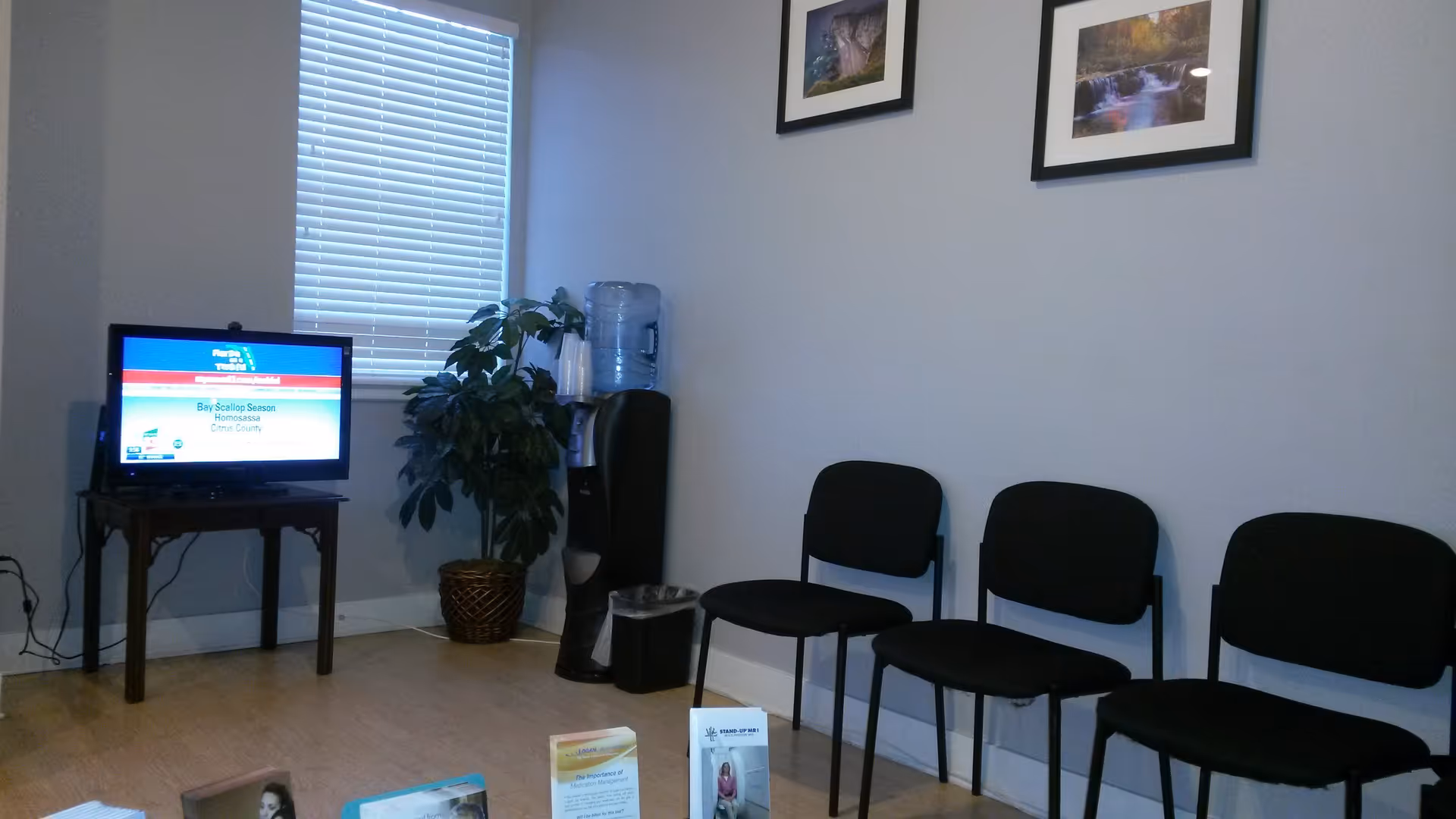 A small indoor waiting area at a senior facility with chairs, a TV, plant, and art.