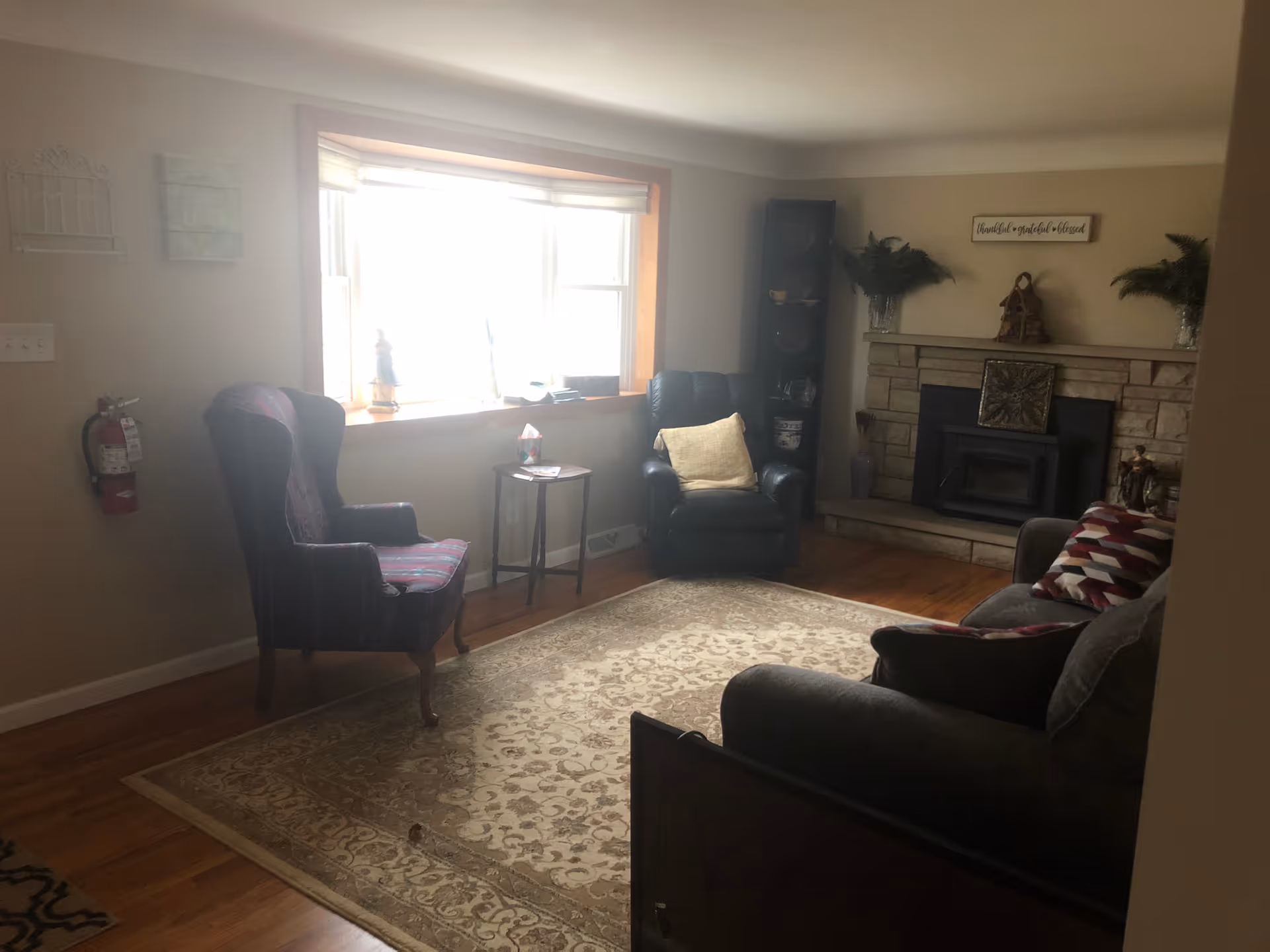 A cozy living room with a large window letting in natural light, a patterned area rug on wooden floor, a dark armchair with a cushion, a striped wingback chair, a small side table, and a stone fireplace decorated with plants and a sign above it.