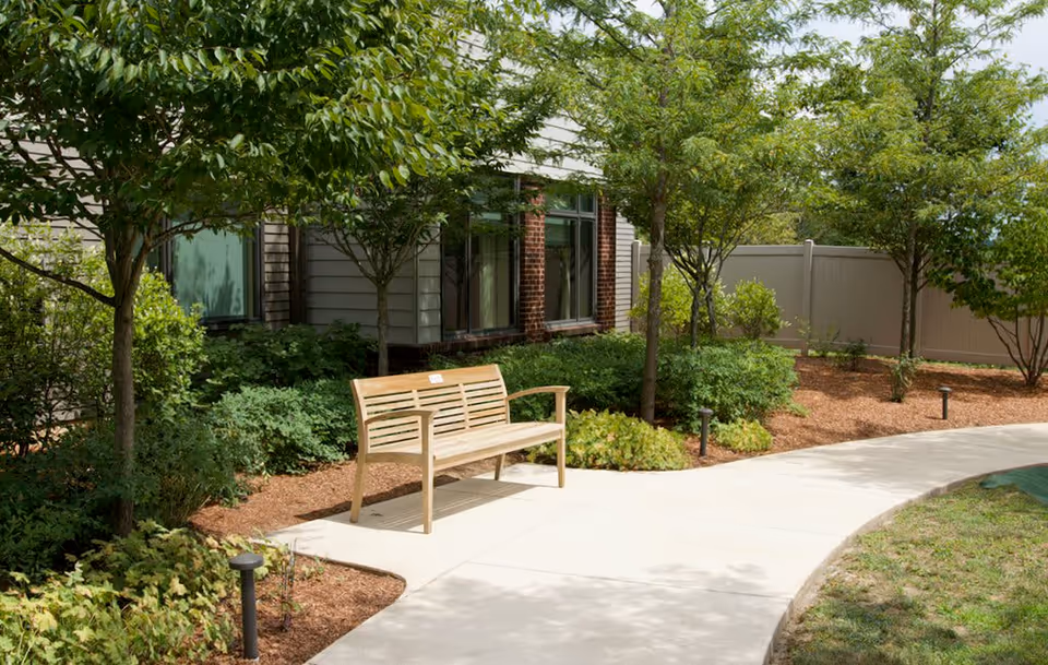 A wooden bench placed on a curved concrete pathway in a garden area with trees, shrubs, and mulch beds next to a building with windows and siding.