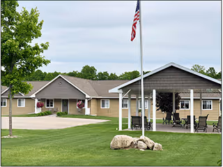 Single-story senior living facility building with a flagpole, covered patio seating, and a manicured lawn.