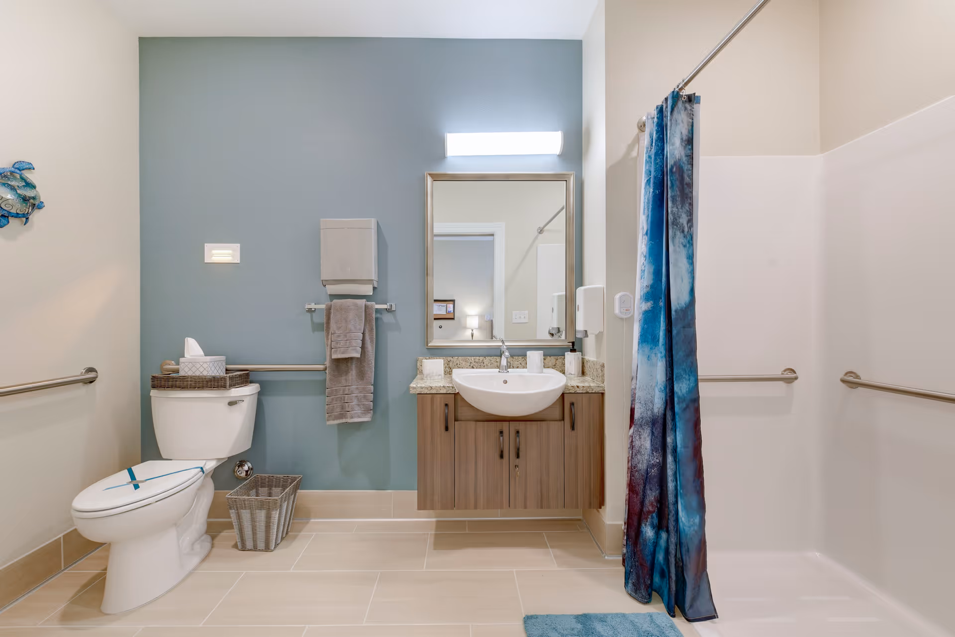 A clean and modern bathroom with a white toilet, a wicker basket, a towel rack with a gray towel, a wall-mounted sink with a granite countertop and wooden cabinet, a rectangular mirror with a light above it, and a shower area with a blue and white patterned shower curtain.