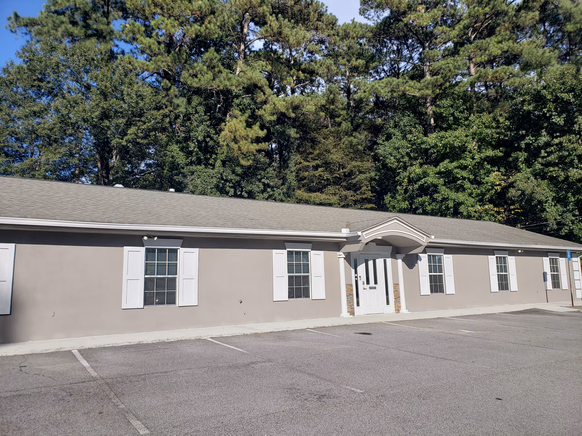 Single-story beige senior care home with white shutters, front entrance, parking lot and trees behind.