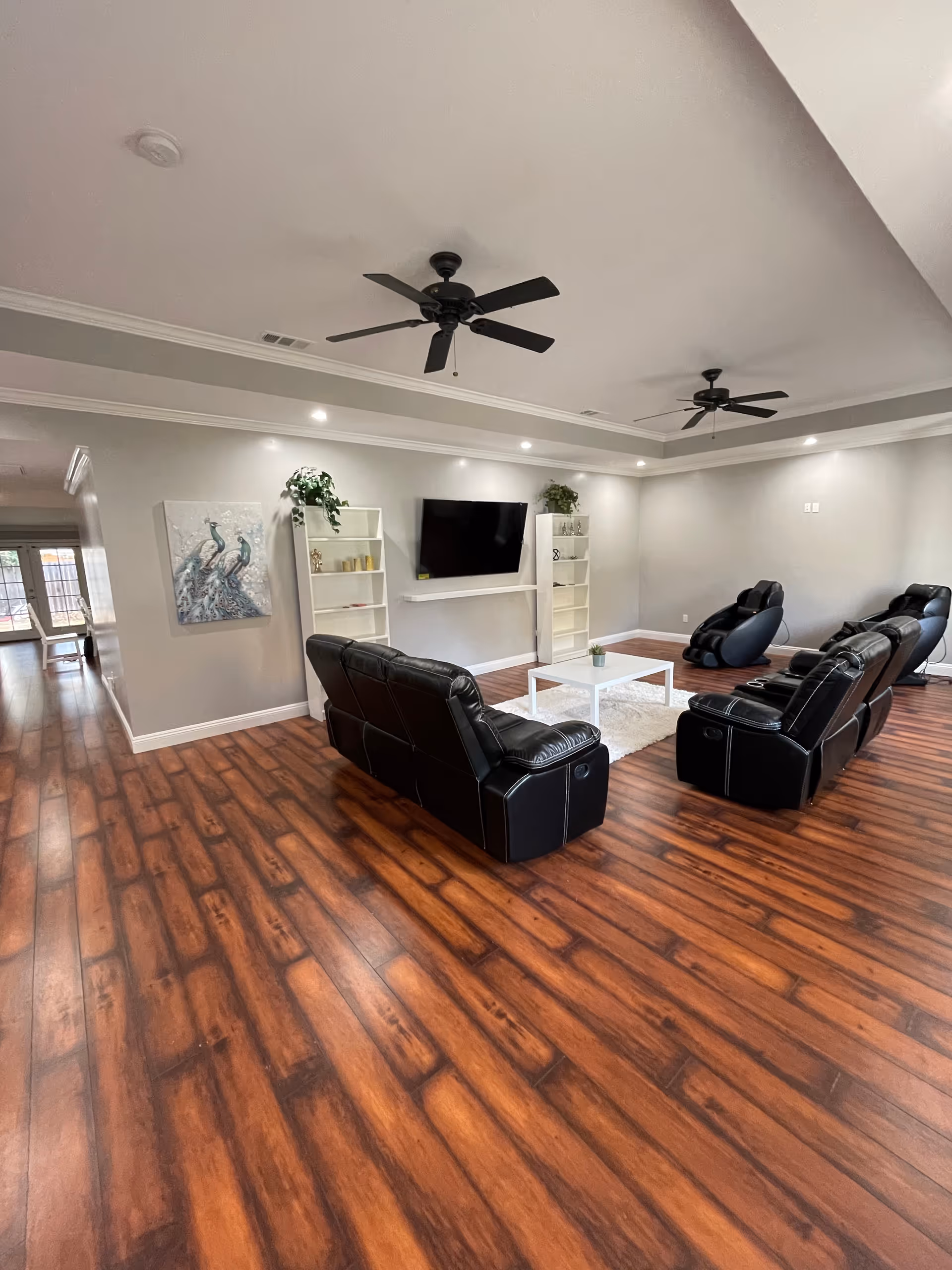Spacious senior living common room with leather recliners and massage chairs facing a wall-mounted TV, white shelving, a coffee table, and ceiling fans.