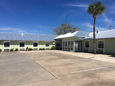 Exterior view of a single-story assisted living facility building with a light green facade, multiple windows, a metal roof, a covered entrance, a tall palm tree, and a large concrete parking area in front under a clear blue sky.