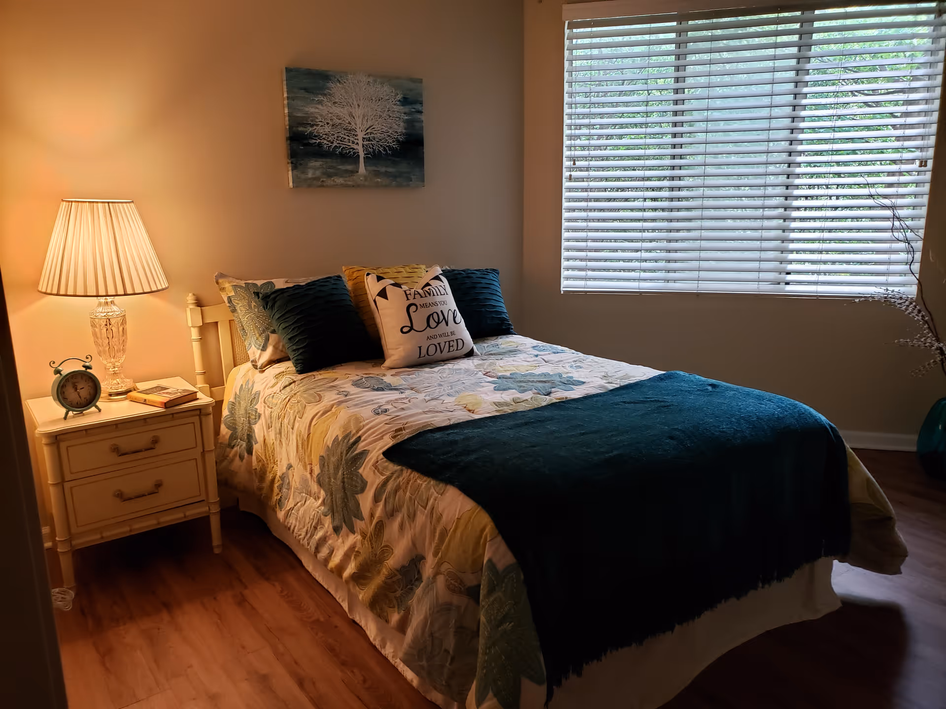 Cozy bedroom with a neatly made bed, bedside table with lamp and alarm clock, and a large window with blinds.