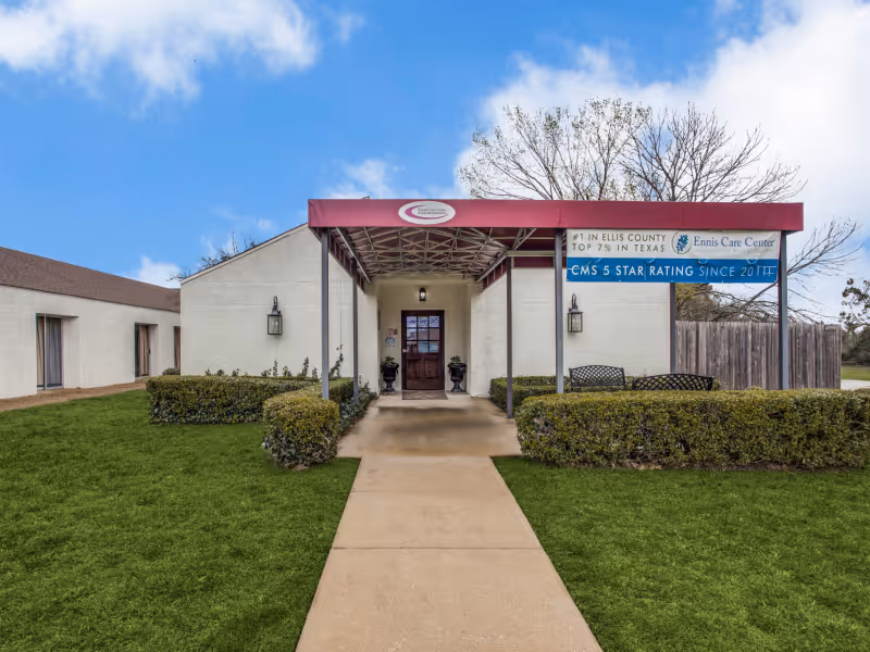 Front entrance of Ennis Care Center with a covered walkway, white exterior walls, green bushes, and a clear blue sky with some clouds.