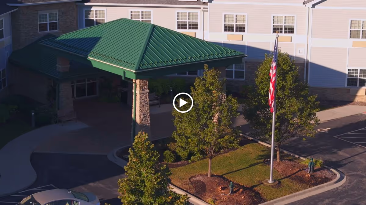 Exterior view of New Perspective Senior Living | Long Grove showing the entrance with a green metal roof canopy, a flagpole with an American flag, several trees, and part of the parking lot.