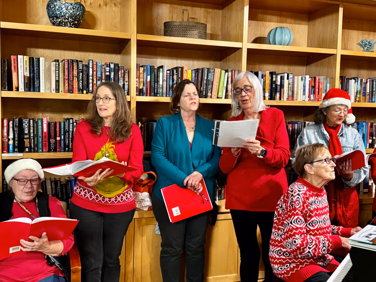 A group of six elderly women standing and sitting in front of wooden bookshelves filled with books. They are holding red songbooks and appear to be singing, with some wearing festive holiday clothing including Santa hats and Christmas sweaters. One woman is seated at a piano.