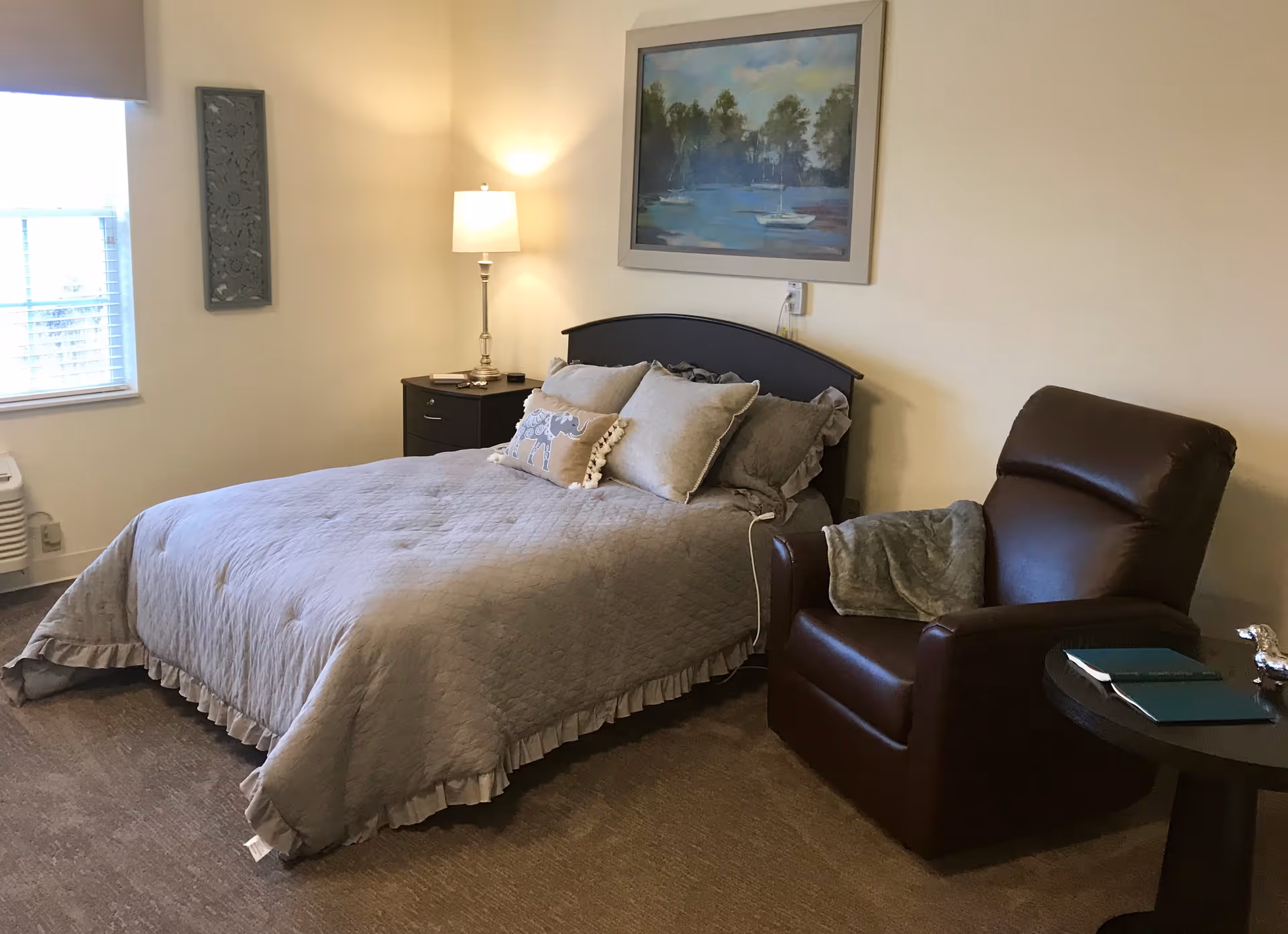 A neatly made bed with a gray quilt and decorative pillows next to a nightstand with a lamp and a brown recliner in a cozy bedroom.