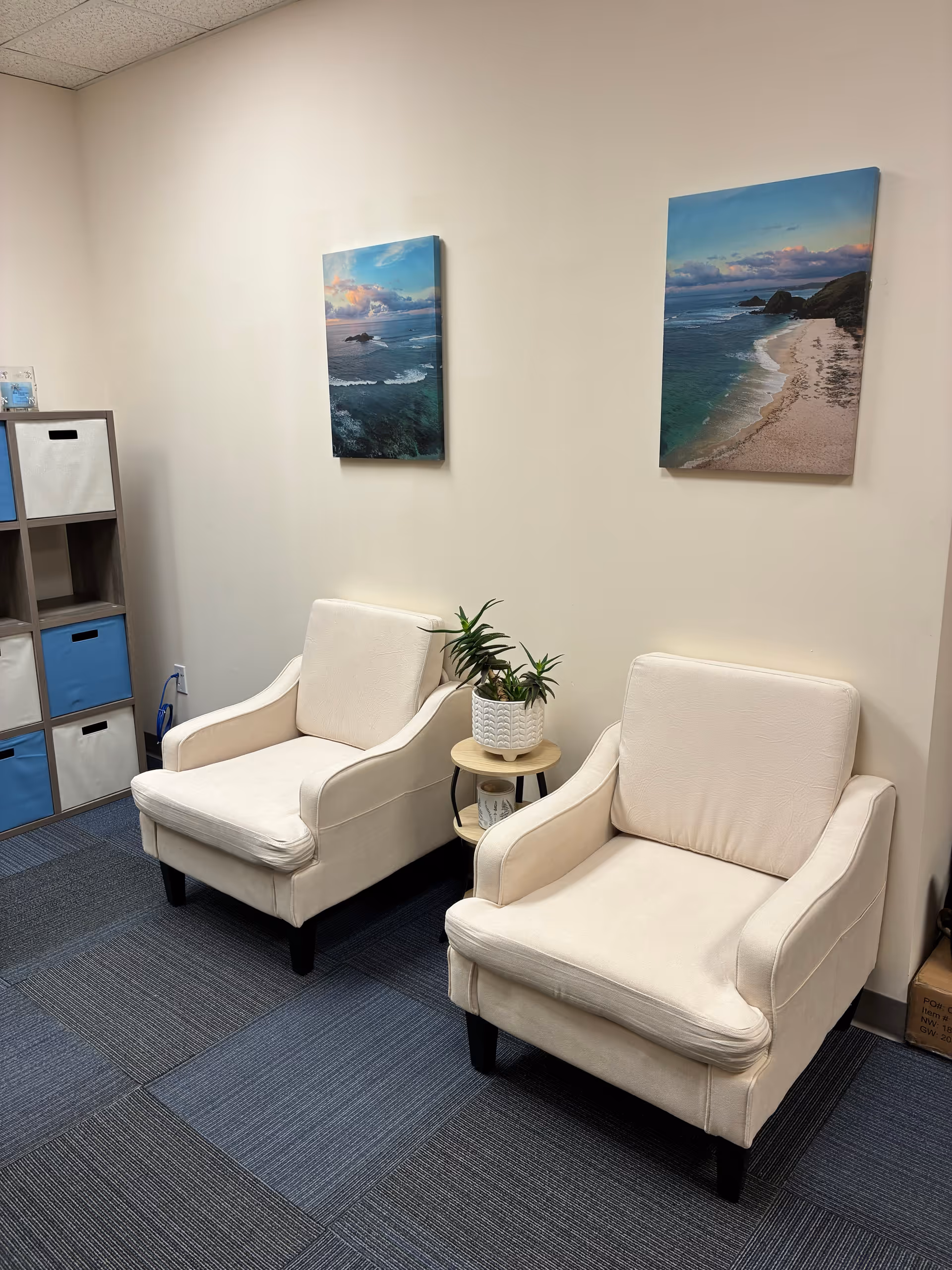 Two white upholstered chairs with a small round table and potted plant in a waiting area.