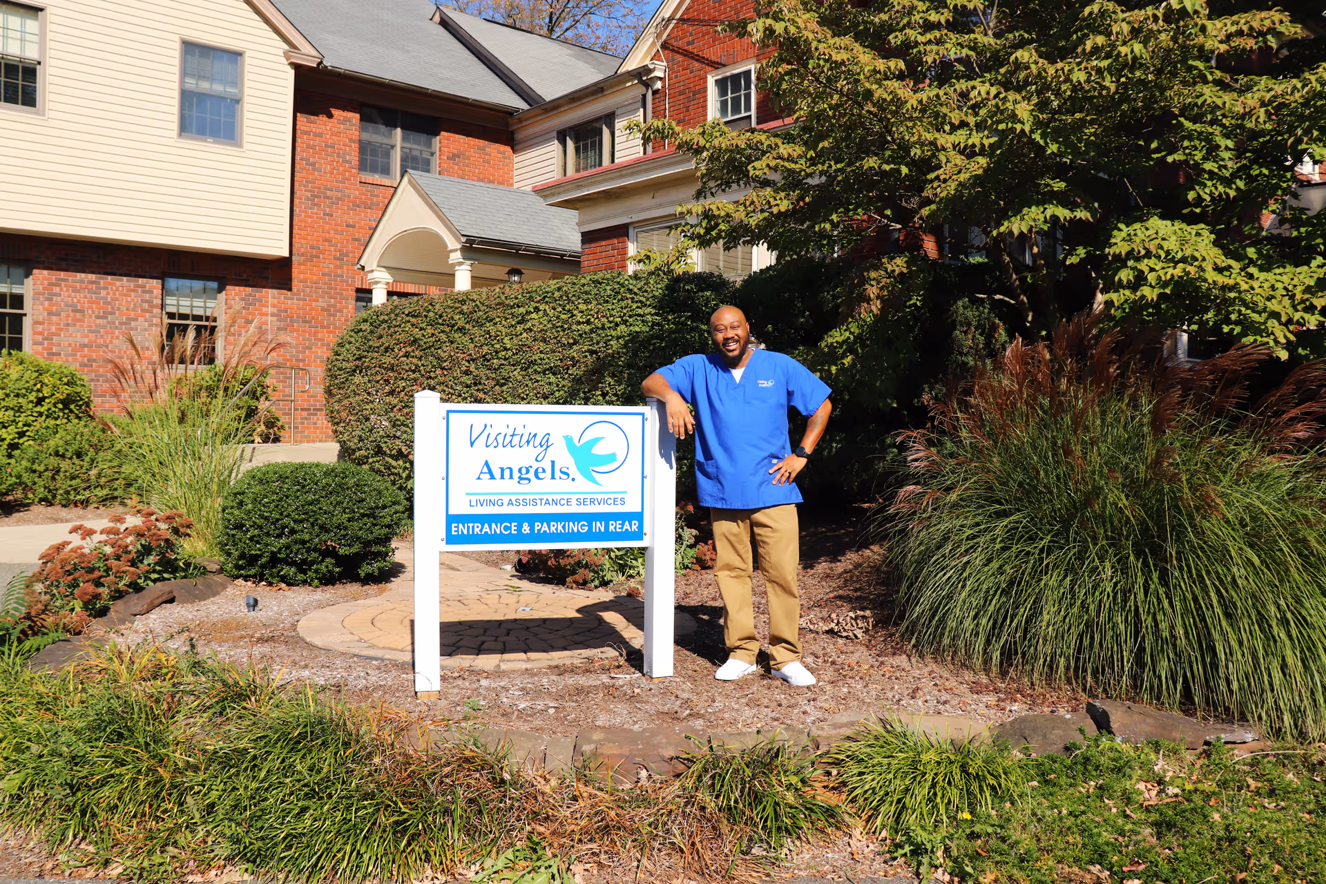 A caregiver in blue scrubs stands beside a Visiting Angels sign in front of brick building.