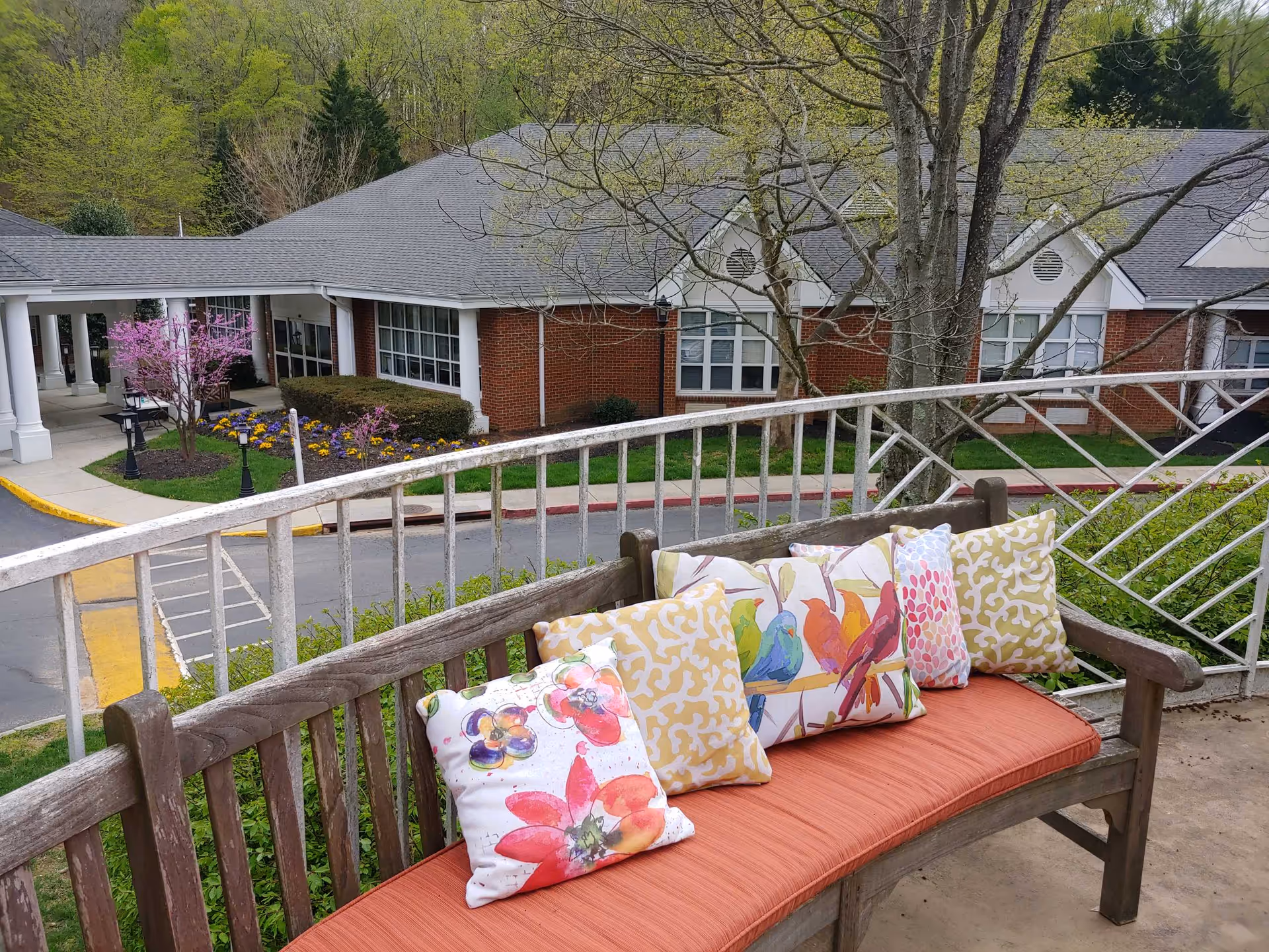 Outdoor wooden bench with an orange cushion and several colorful decorative pillows featuring floral and bird designs, situated on a balcony overlooking the entrance of a brick building with white columns, surrounded by trees and flower beds.