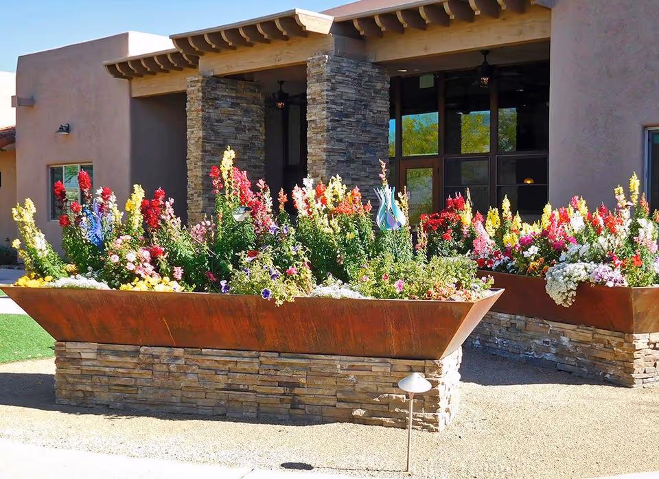 Two large rectangular flower beds filled with colorful blooming flowers in front of a building entrance with stone pillars and wooden beams under a clear blue sky.