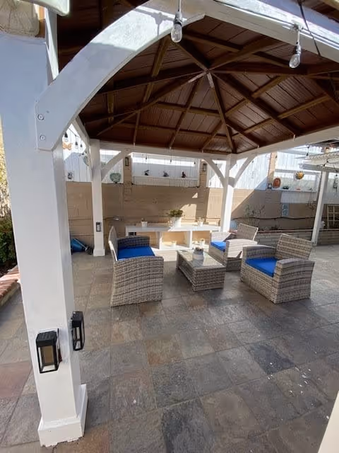 Outdoor covered seating area with a wooden roof structure, four wicker chairs with blue cushions arranged around a glass-top coffee table, and a white bench against a beige wall with small decorative items and plants.