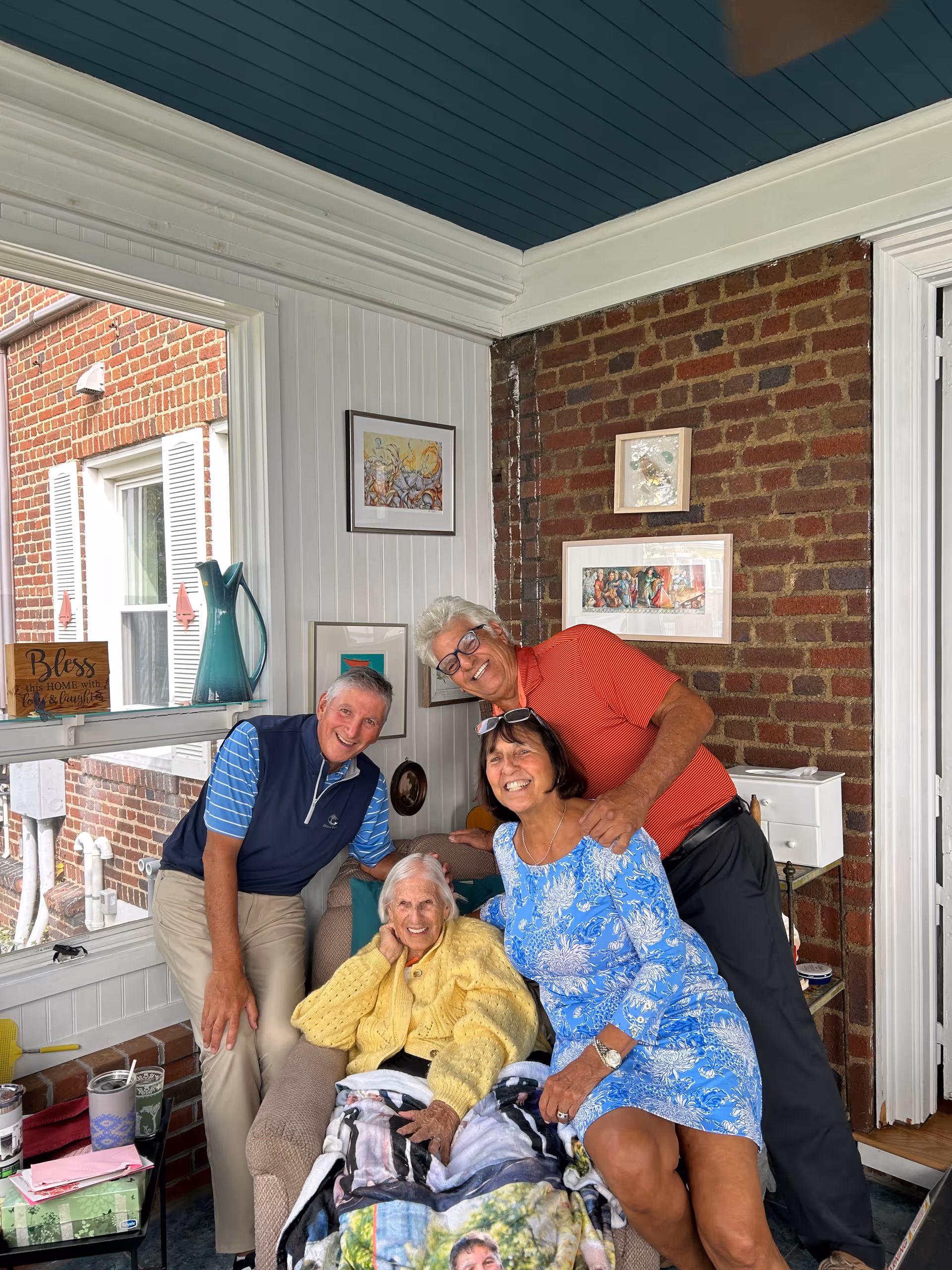 Group of four adults posing with an elderly woman on a sunlit outdoor porch.