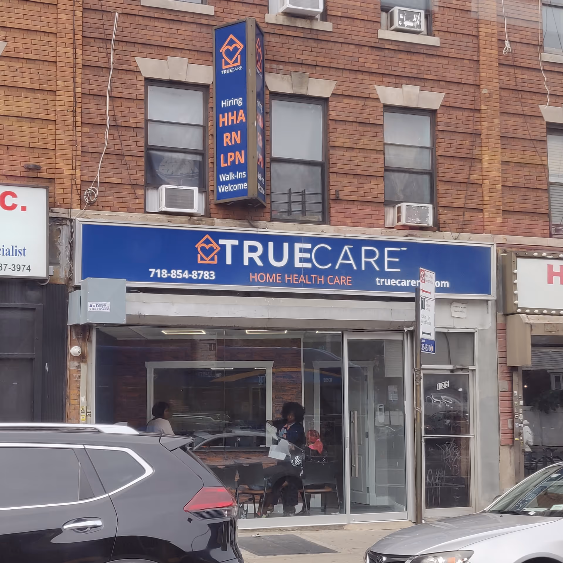 Brick storefront with a blue TrueCare sign and people seated near the glass entrance.