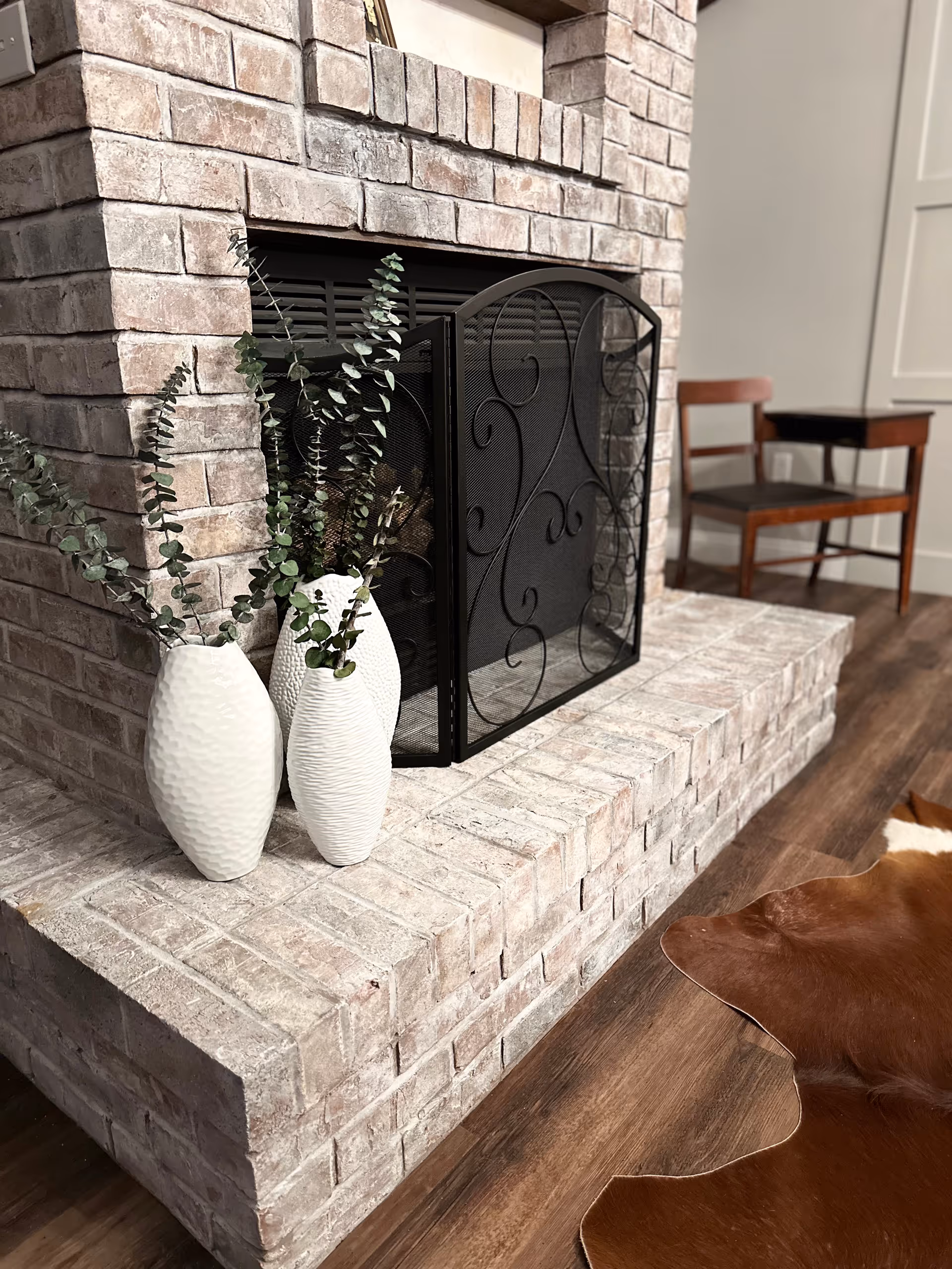 Brick fireplace with decorative vases on the hearth, chairs, wood floor, and cowhide rug.