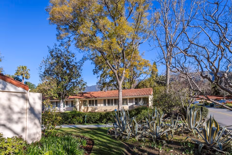 Single-story building with a red tile roof set behind landscaped gardens, agave plants, and trees under a clear blue sky.