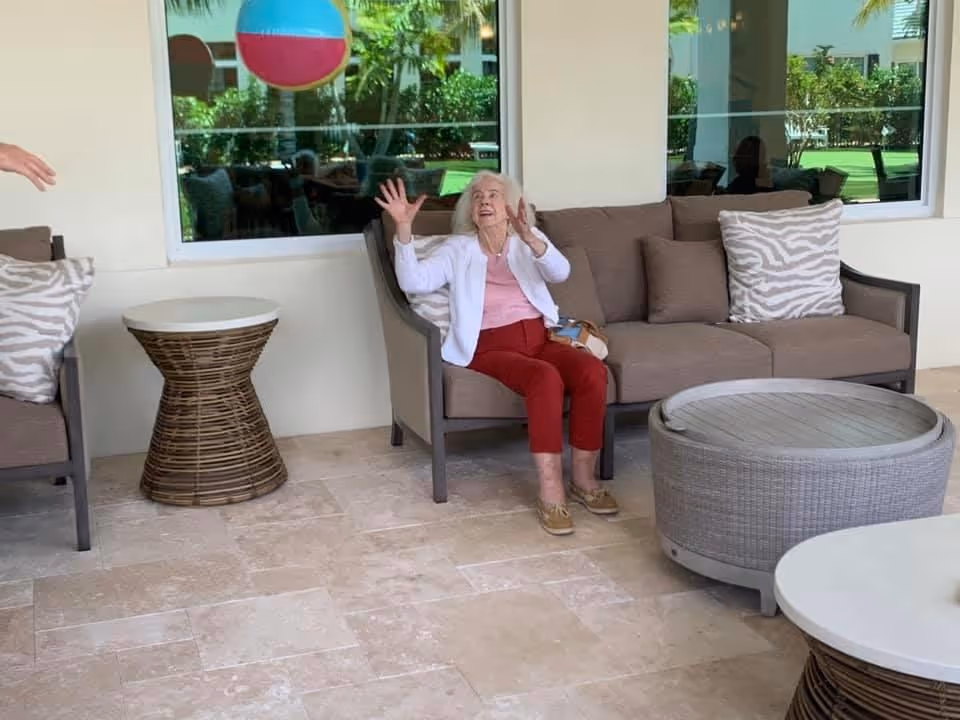 An elderly woman sitting on a cushioned outdoor sofa under a covered patio, reaching up to catch a colorful beach ball. The patio has tiled flooring, wicker side tables, and large windows showing greenery outside.