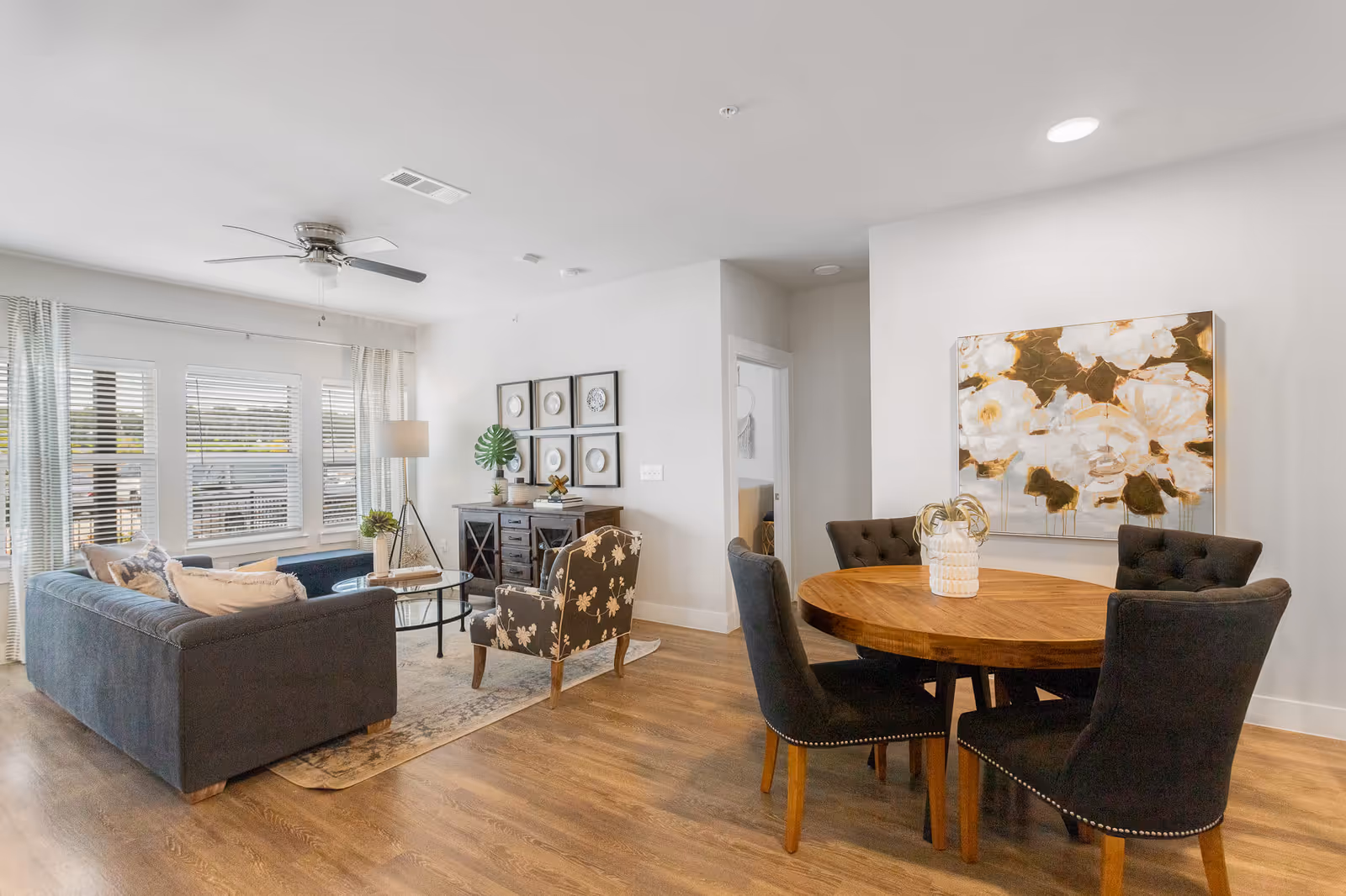 A bright and modern living and dining area with wooden flooring. The living room features a dark gray sofa, a floral patterned armchair, a glass coffee table, a floor lamp, and a sideboard with decorative items. The dining area has a round wooden table with four dark upholstered chairs and a large floral painting on the wall.