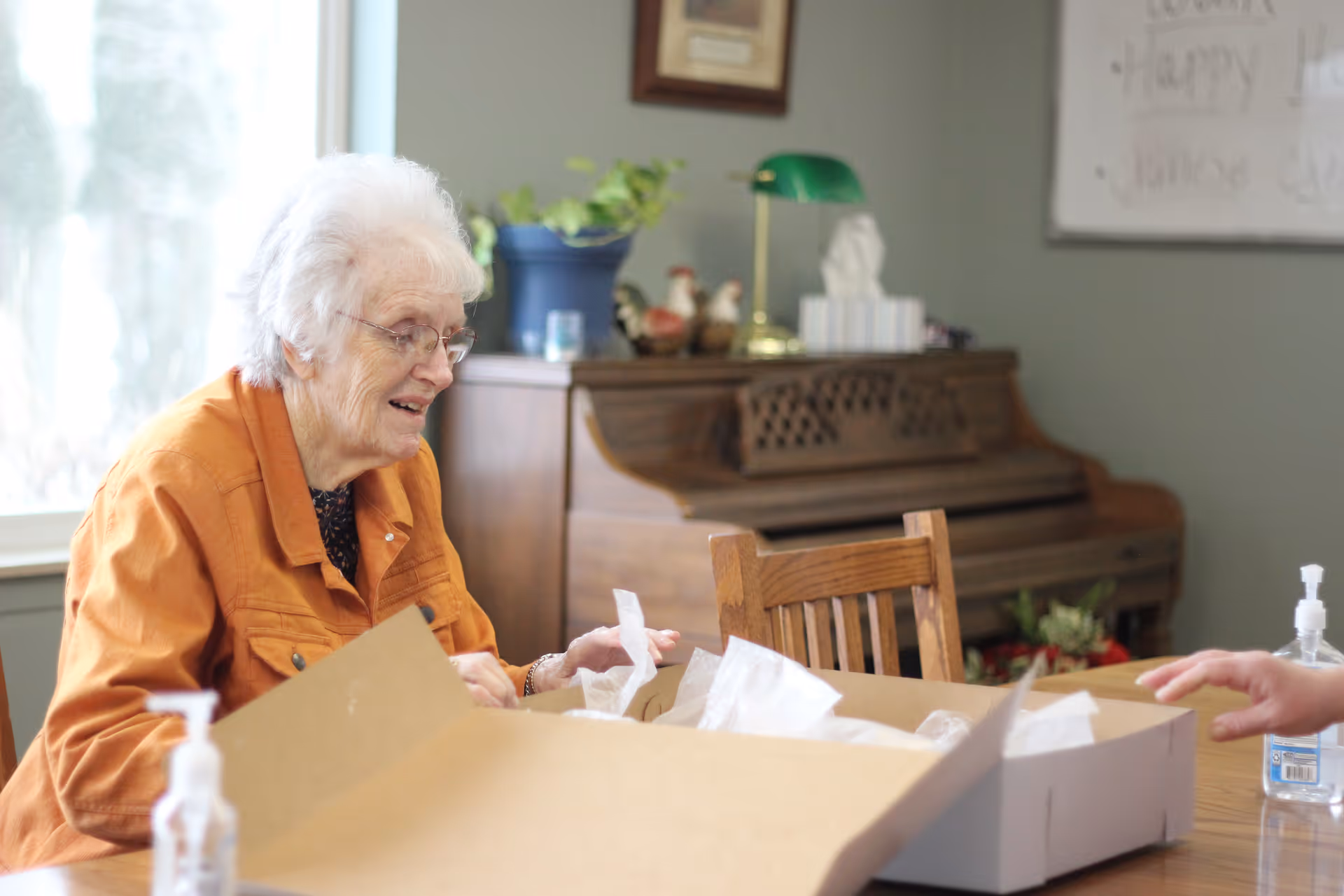 An elderly woman with white hair and glasses, wearing an orange jacket, is sitting at a wooden table and smiling while unpacking a large cardboard box. In the background, there is a wooden piano with decorative items on top, a green desk lamp, and a potted plant. A hand of another person is visible on the right side of the table. There are also two bottles of hand sanitizer on the table.