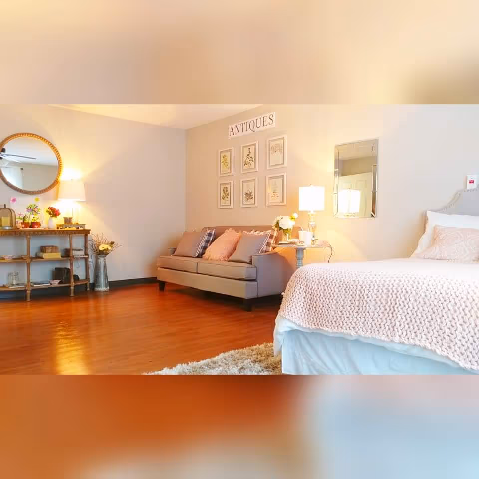 A cozy bedroom with a neatly made bed covered in a light pink knitted blanket and pillows. To the right of the bed is a small round side table with a lamp and flowers. Against the wall is a gray sofa with several cushions. Above the sofa are six framed botanical prints and a sign that reads 'ANTIQUES'. On the left side of the room is a wooden console table with decorative items and a round mirror hanging above it. The room has warm wooden flooring and soft lighting.