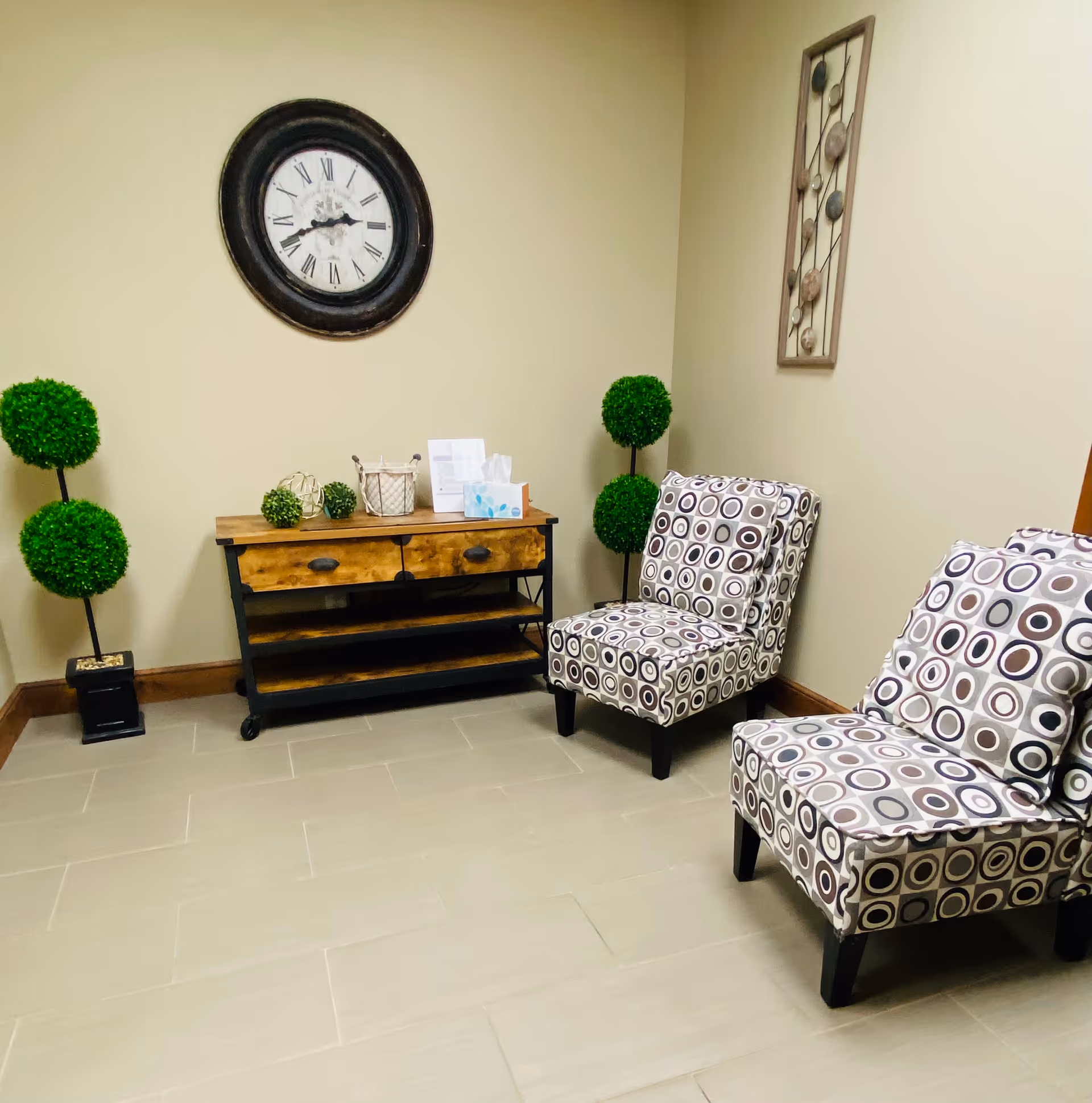 Neutral lounge area with patterned chairs, a wooden console, clock, and decorative topiaries.