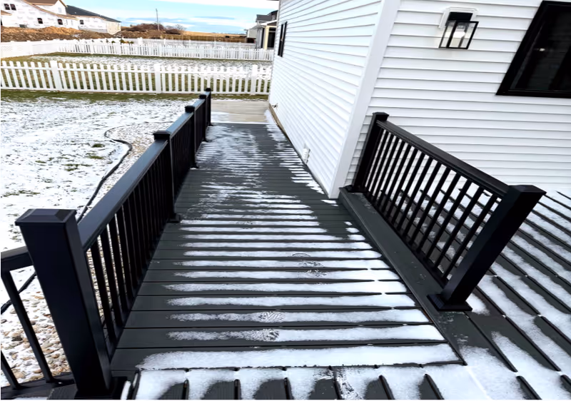 Snowy exterior ramp with black railing beside a white house and fenced yard.