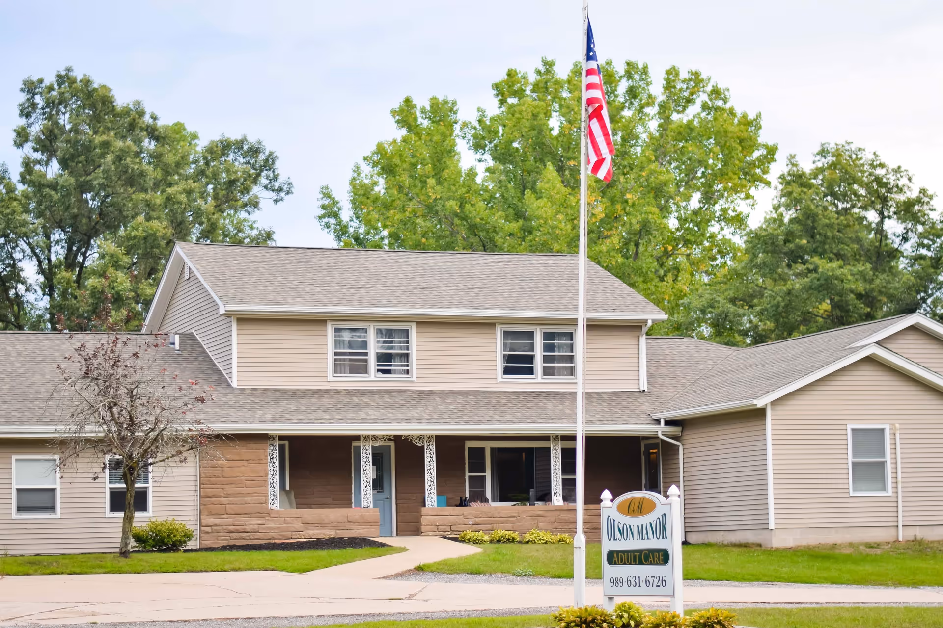 Exterior view of Olson Manor, a two-story beige building with a front porch, surrounded by green trees and grass. An American flag is flying on a flagpole in front of the building, and a sign in the lawn reads 'Olson Manor Adult Care' with a phone number.
