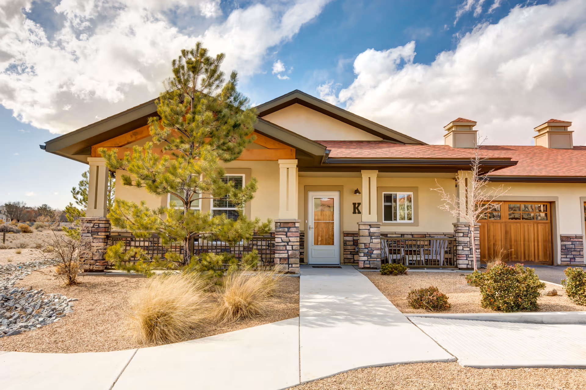 Single-story ranch-style building front with a covered porch, garage, drought-tolerant landscaping, and a tree under a partly cloudy sky.