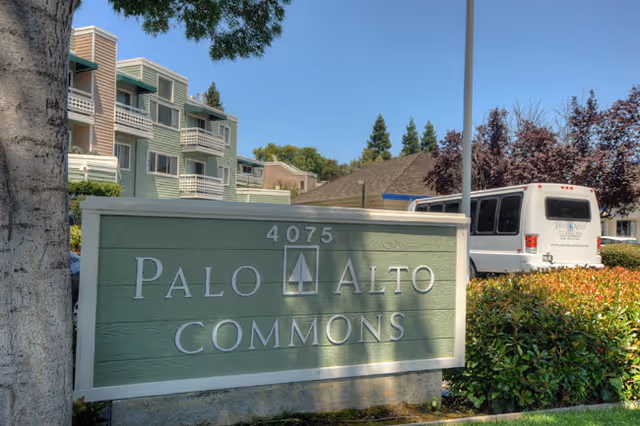 Entrance sign reading "Palo Alto Commons" with the address 4075 in front of residential buildings, landscaping, and a parked shuttle bus.