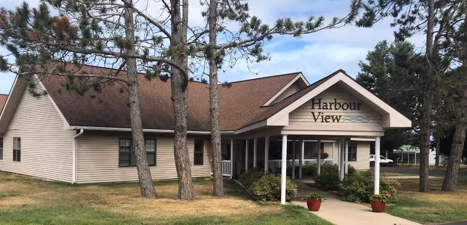 Front entrance of a single-story beige building with a covered porch and a sign reading 'Harbour View', surrounded by trees and lawn.