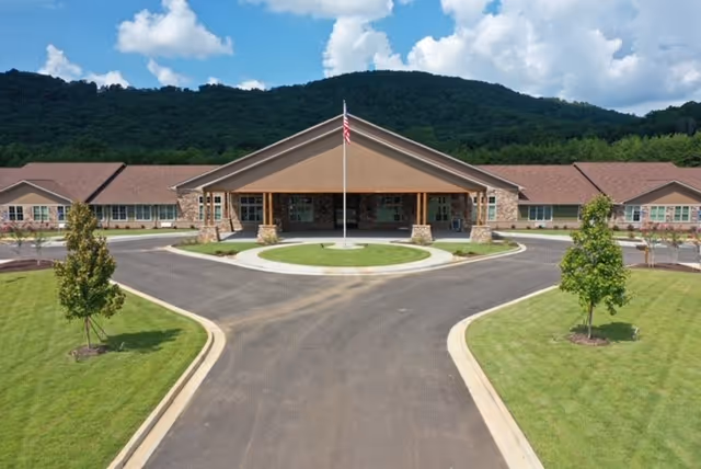 Front entrance of a single-story assisted living building with a circular driveway, central flagpole, and landscaped lawns set against wooded hills.