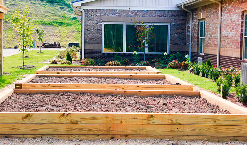 Raised garden beds filled with soil in front of a brick building with windows, surrounded by small trees and shrubs on a sunny day.