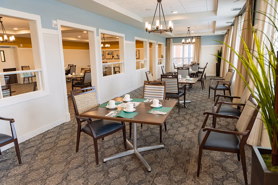 A bright dining room in a senior living facility with several wooden tables and chairs arranged neatly. Each table is set with white cups, saucers, and silverware on green placemats. Large windows with beige curtains allow natural light to fill the room. There are decorative plants near the windows and modern chandeliers hanging from the ceiling. The walls are painted light blue and white, and there are interior windows looking into an adjacent room.
