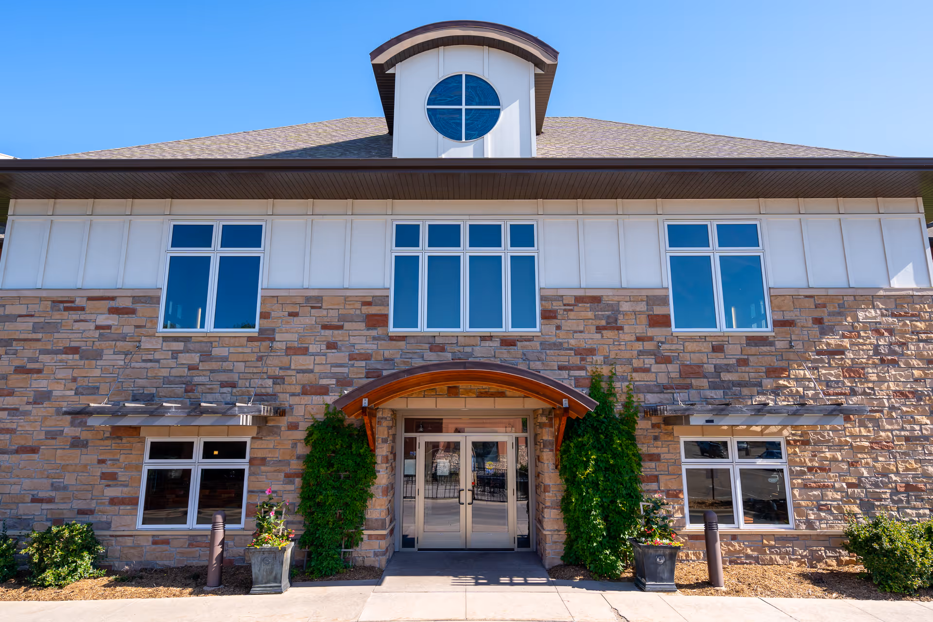 Front exterior view of a two-story building with stone and siding facade, large windows, a curved wooden awning over the entrance, and greenery on either side of the doorway under a clear blue sky.