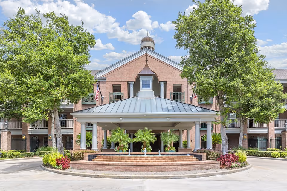 Front exterior view of a senior living facility named Brookdale West University, featuring a large covered entrance with columns, brick facade, balconies, and landscaped greenery including trees and shrubs under a partly cloudy sky.