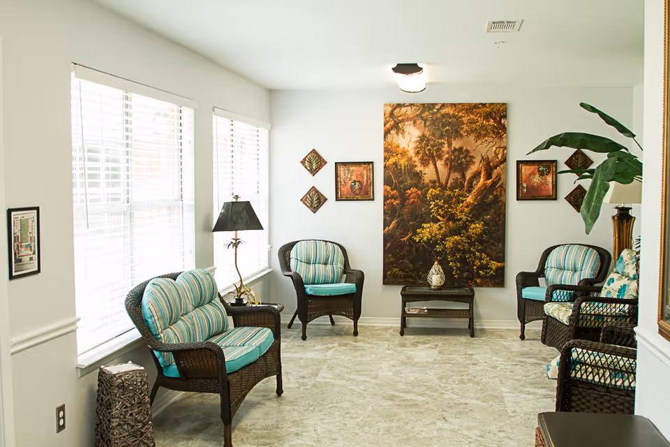 Bright communal seating area with wicker chairs with striped cushions, a central table and large landscape artwork on the back wall.