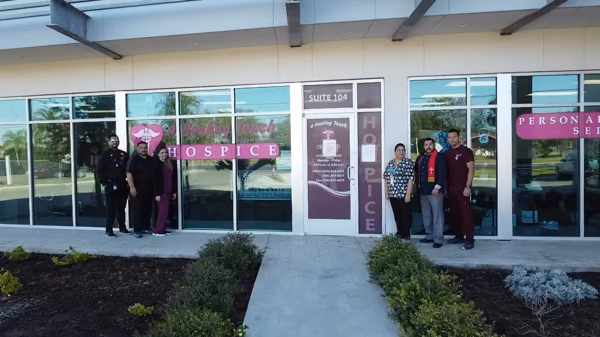 Six people stand outside a hospice entrance with pink signage and glass storefront.