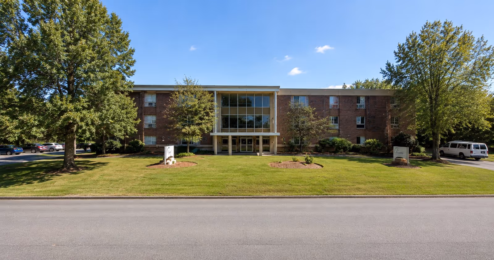 Brick senior living center with glass-front entrance, well-kept lawn, and surrounding trees.